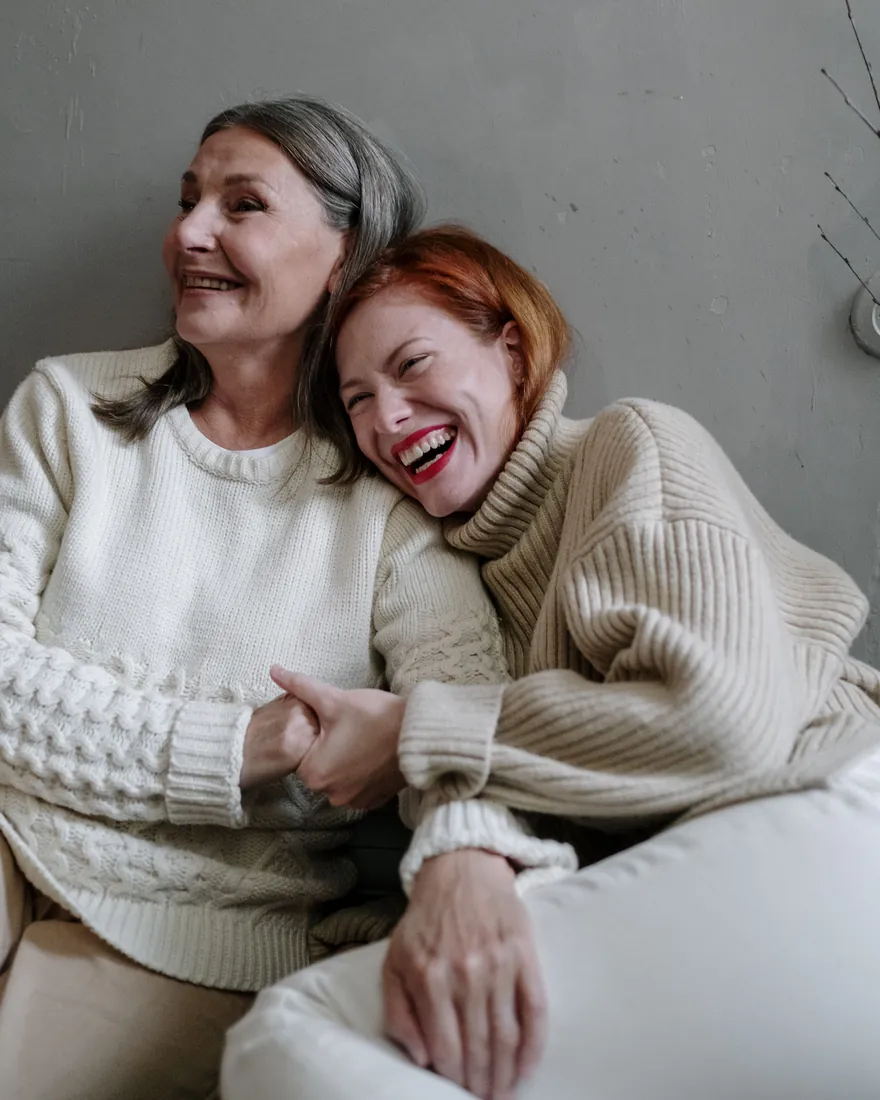 An older woman and a younger woman laughing together, leaning on each other in cream knit sweaters